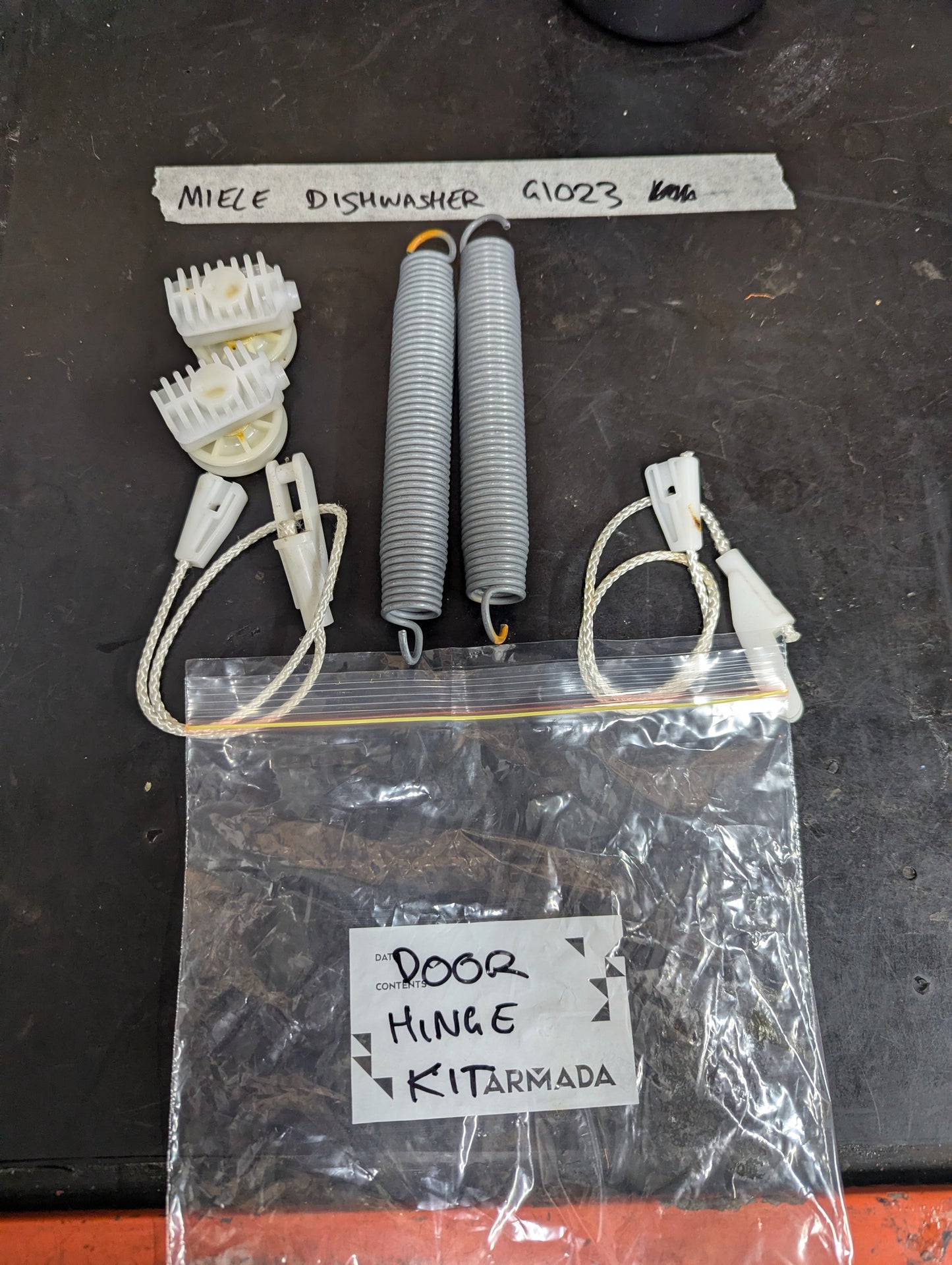 Miele G1023 dishwasher door hinge kit with springs, cables, and pulleys displayed on a black surface