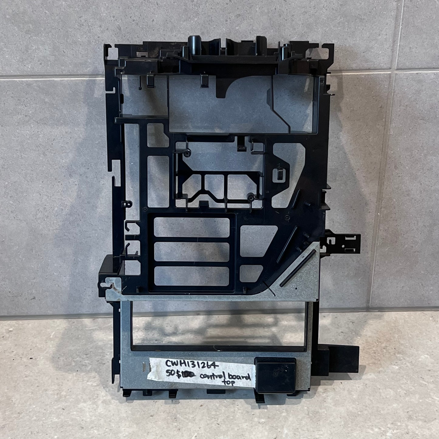 Control board top frame assembly with black plastic and metal reinforcement, labeled for identification on a tile background