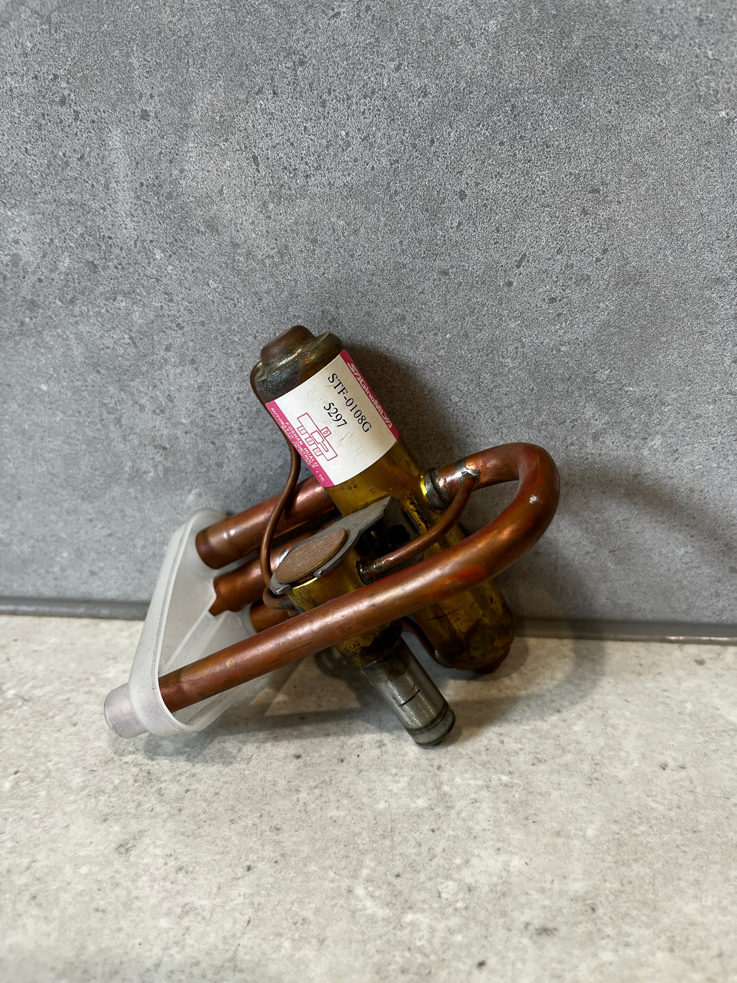 Side view of copper four-way reversing valve with attached metal parts and label, placed on gray surface.