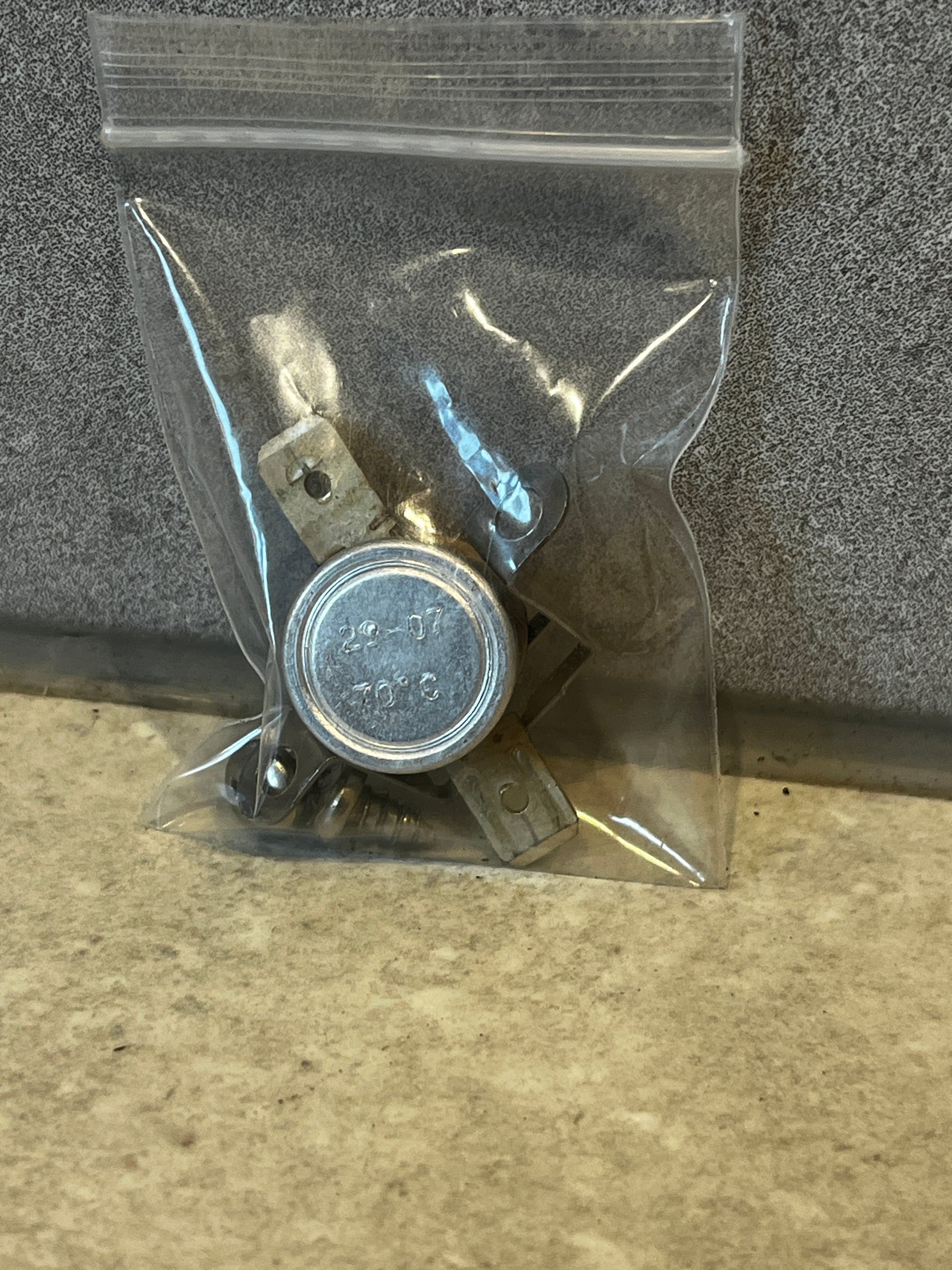 Metal thermostat temperature switch with mounting tabs inside a small clear plastic bag