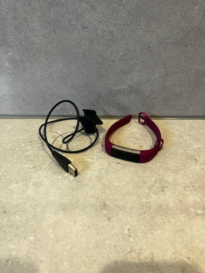 Smart fitness tracker with magenta band and USB charging cable on a stone surface
