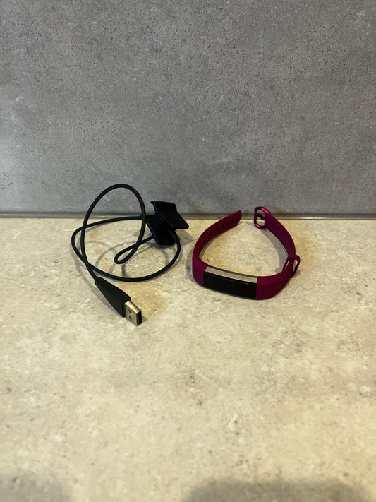 Smart fitness tracker with magenta band and USB charging cable on a stone surface