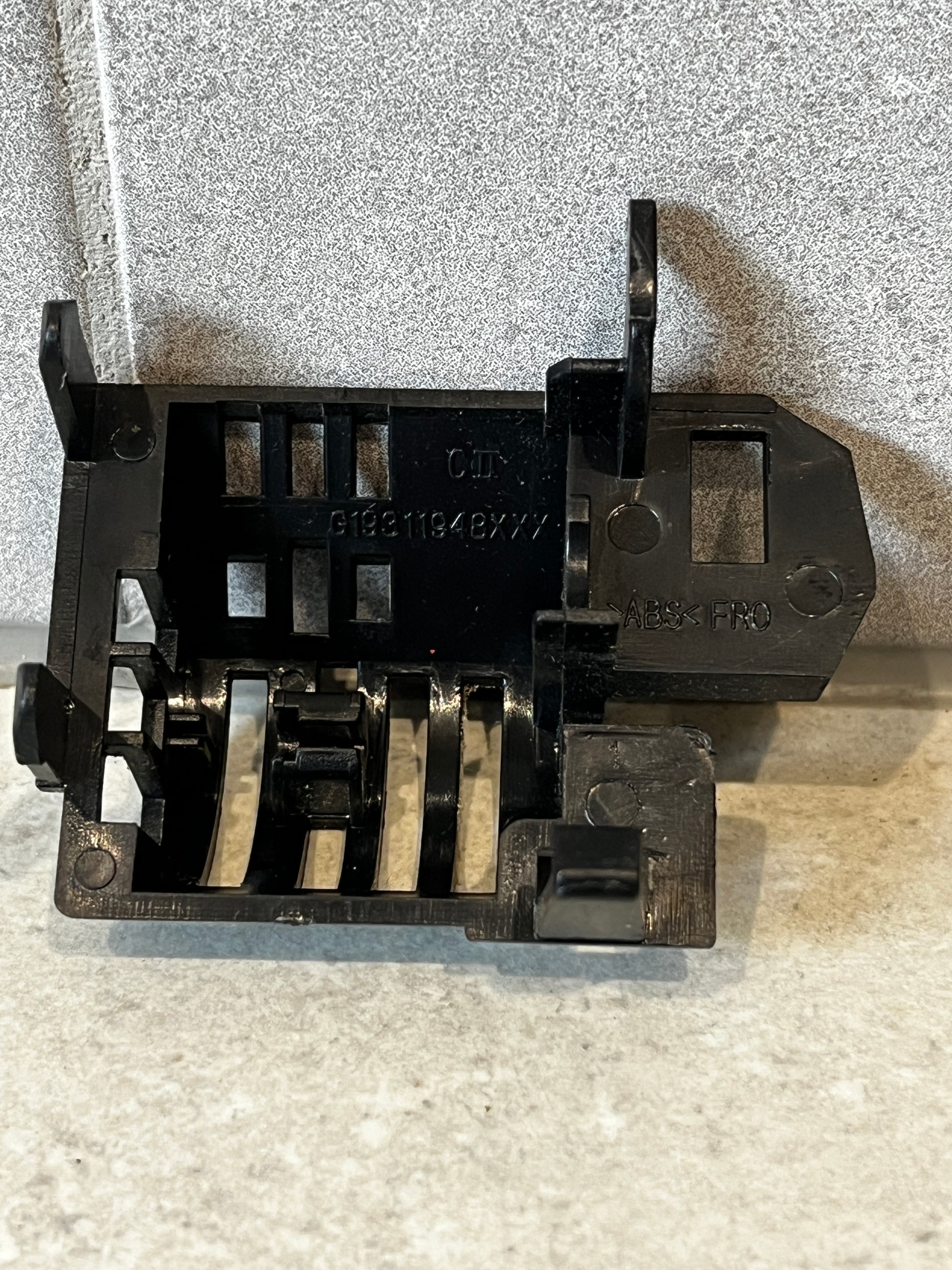 Black ABS plastic mounting bracket with multiple slots and ridges, positioned on a stone surface