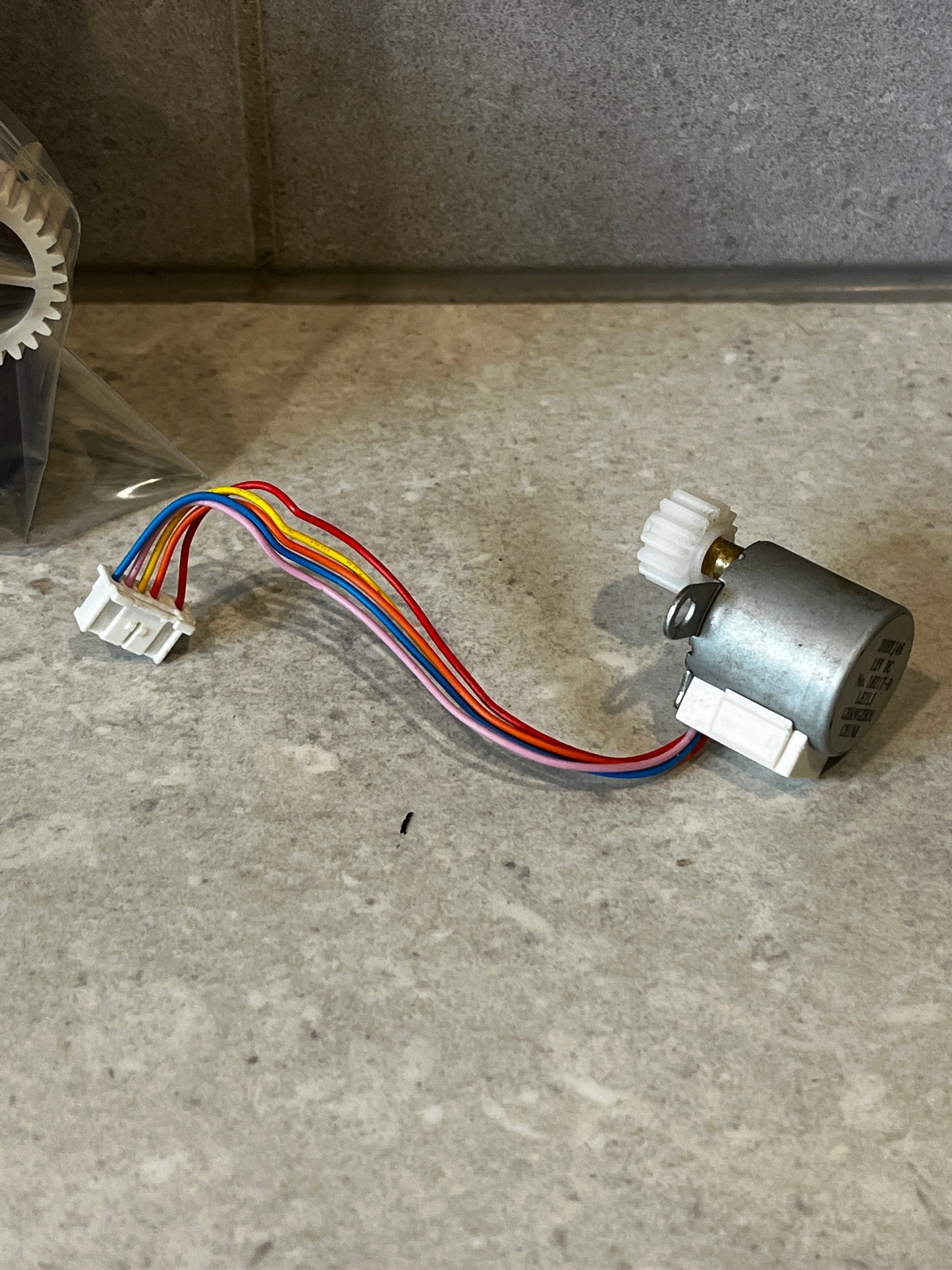 12V DC stepper motor with connected colored wires and white gear on a stone surface