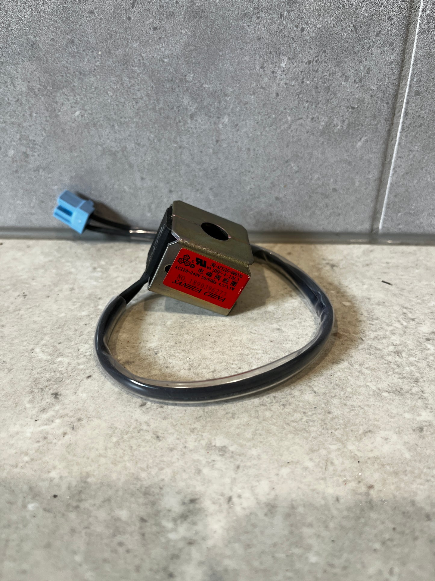 Industrial solenoid coil with metal housing and blue connector on gray tile background