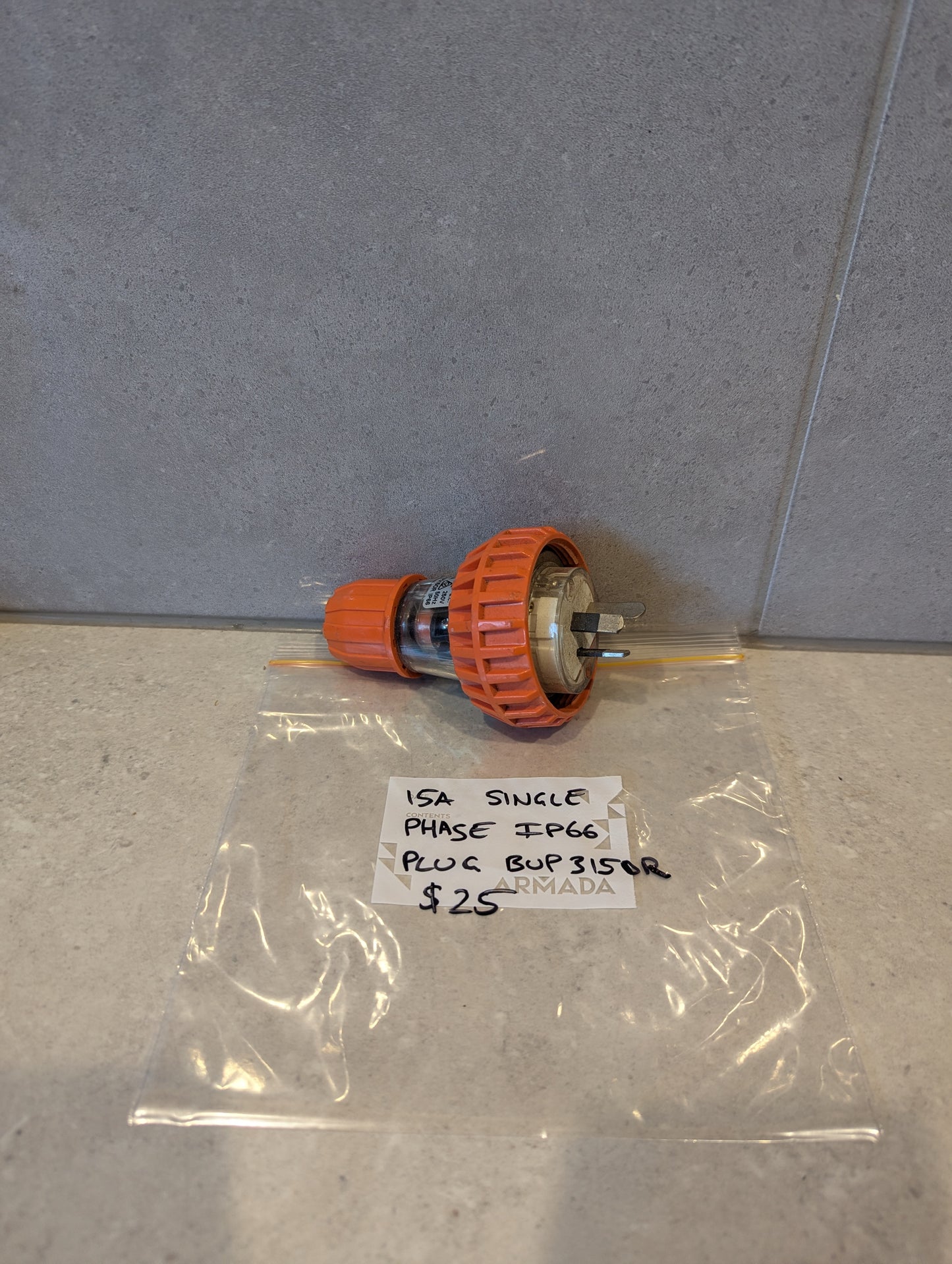 15A Single Phase IP66 electrical plug with orange locking collar and visible prongs, displayed on a counter next to labeled bag