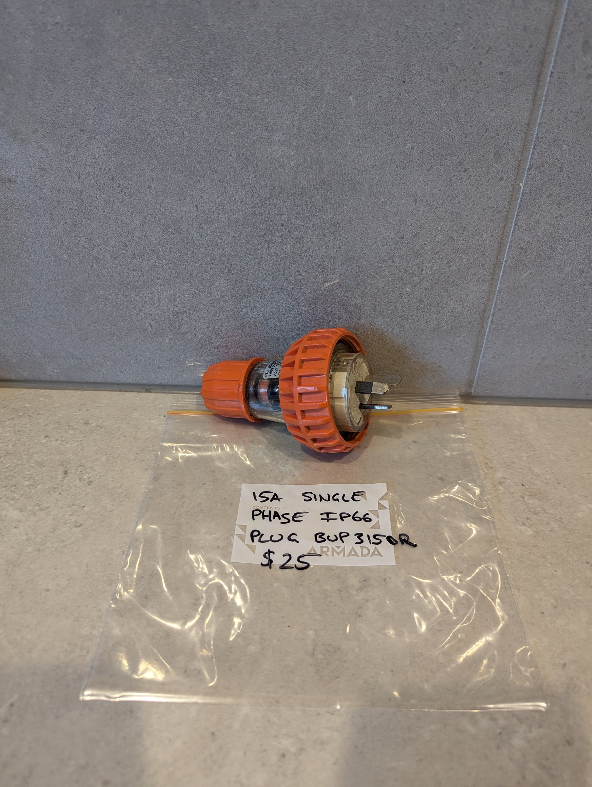 15A Single Phase IP66 electrical plug with orange locking collar and visible prongs, displayed on a counter next to labeled bag