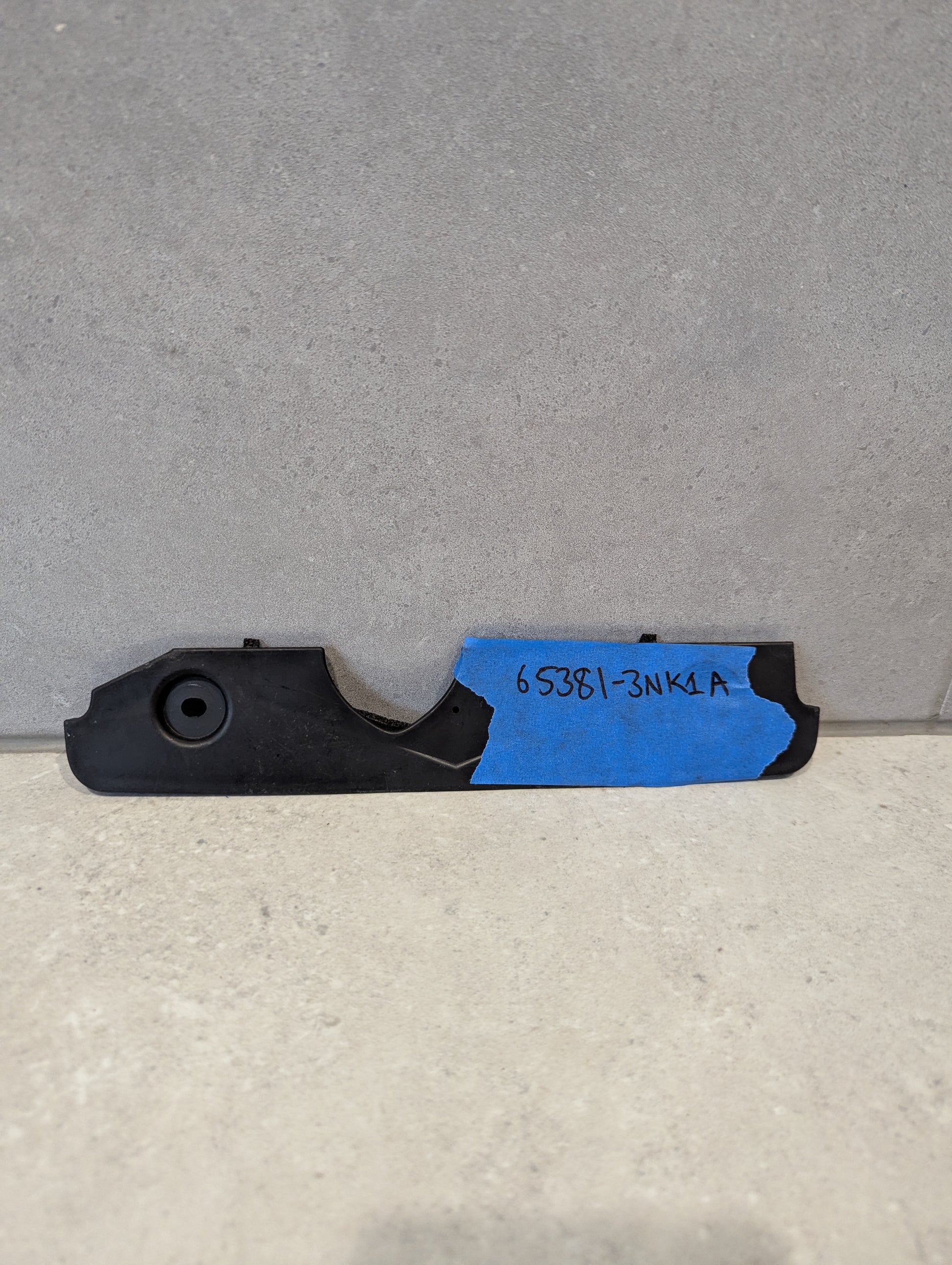 Black plastic automotive engine cover bracket with blue tape and handwritten part number 65381-3NK1A
