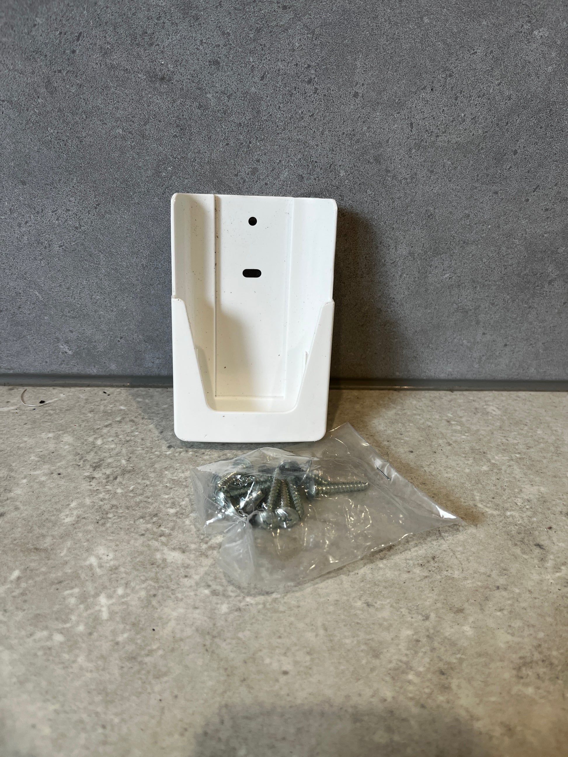 White plastic wall mount bracket with a bag of screws on a gray stone surface
