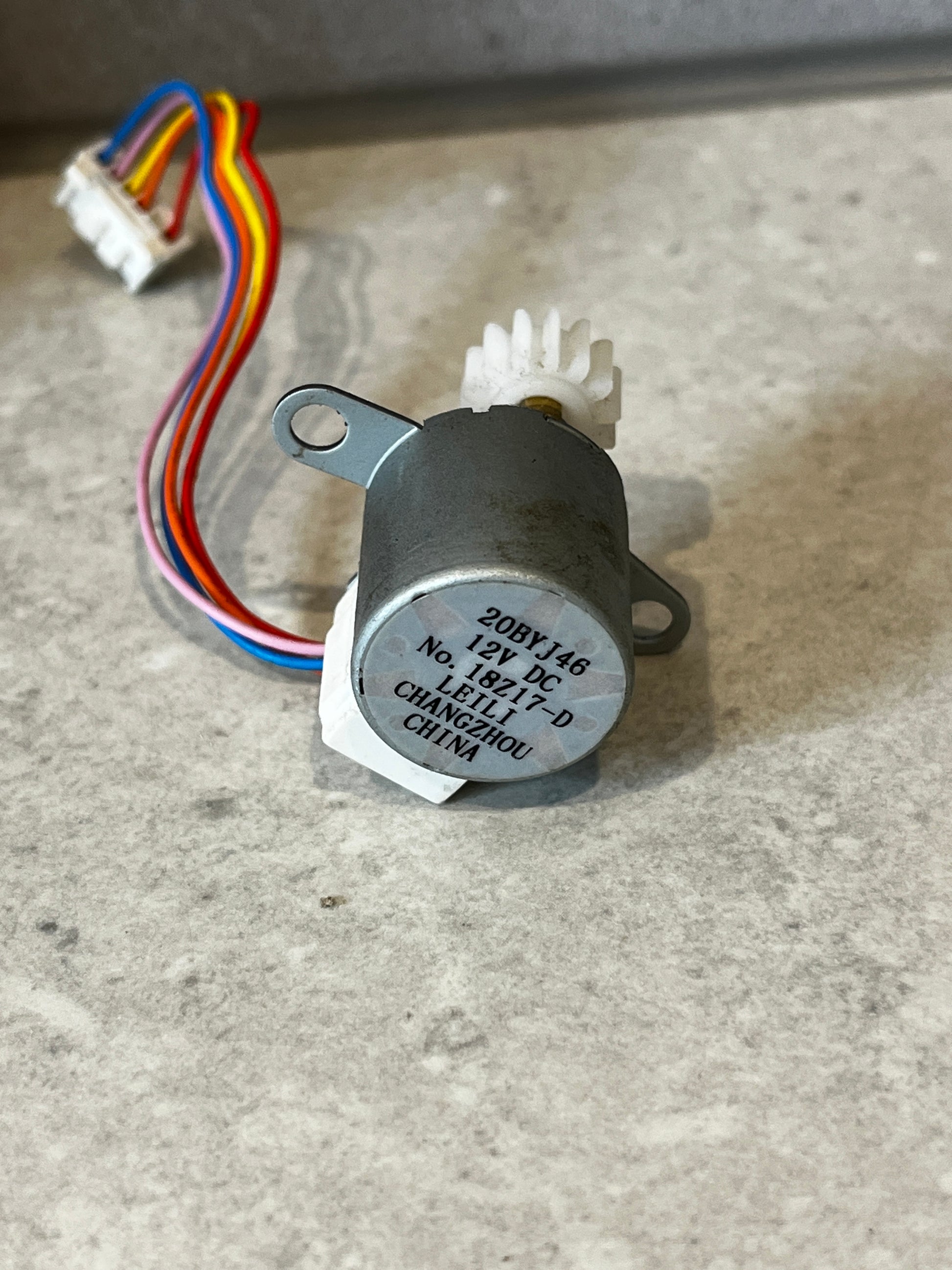 Close-up of 12V DC stepper motor with manufacturing label, colored wires, and white gear