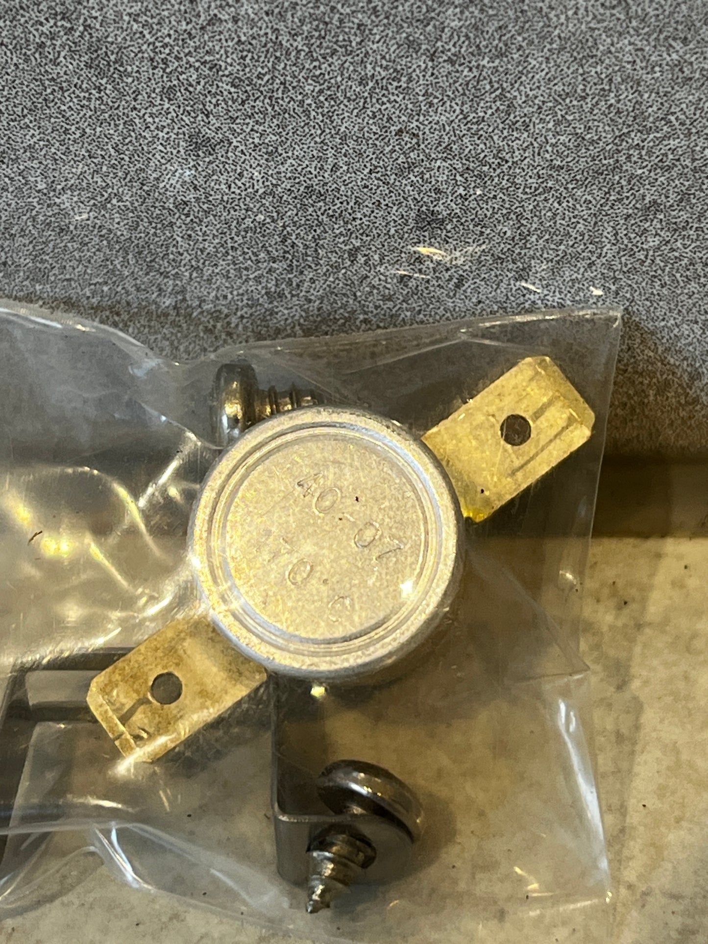 Close-up of metal thermal switch showing brass terminals and mounting bracket inside plastic bag